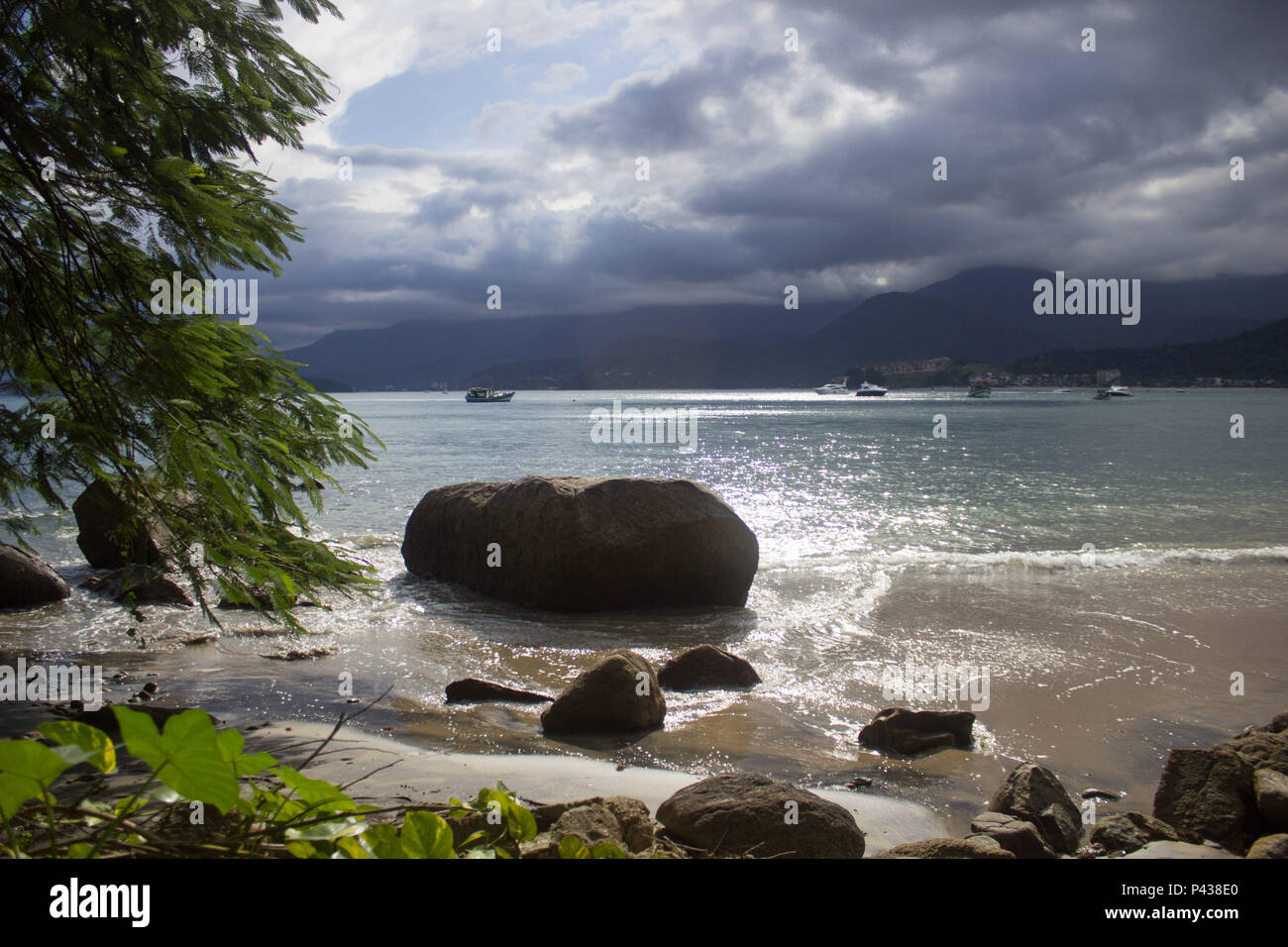 Rochas e pedras em praia ensolarada com barcos navegando no mar ao longe em Ubatuba, SP, Brasil. Stock Photo