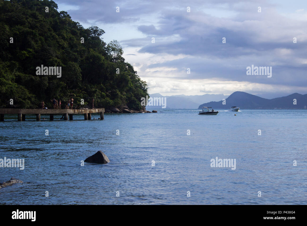 Pessoas sobre pier em costa arborizada com rochas e pedras a beira mar  com barcos navegando e com montanhas ao fundo na praia de Ubatuba, SP, Brasil Stock Photo