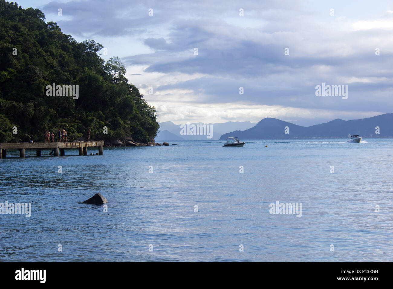 Pessoas sobre pier em costa arborizada com rochas e pedras a beira mar  com barcos navegando e com montanhas ao fundo na praia de Ubatuba, SP, Brasil Stock Photo