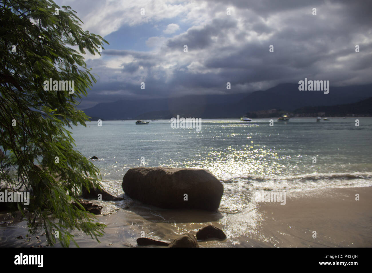 Rochas e pedras em praia ensolarada com barcos navegando no mar ao longe em Ubatuba, SP, Brasil. Stock Photo