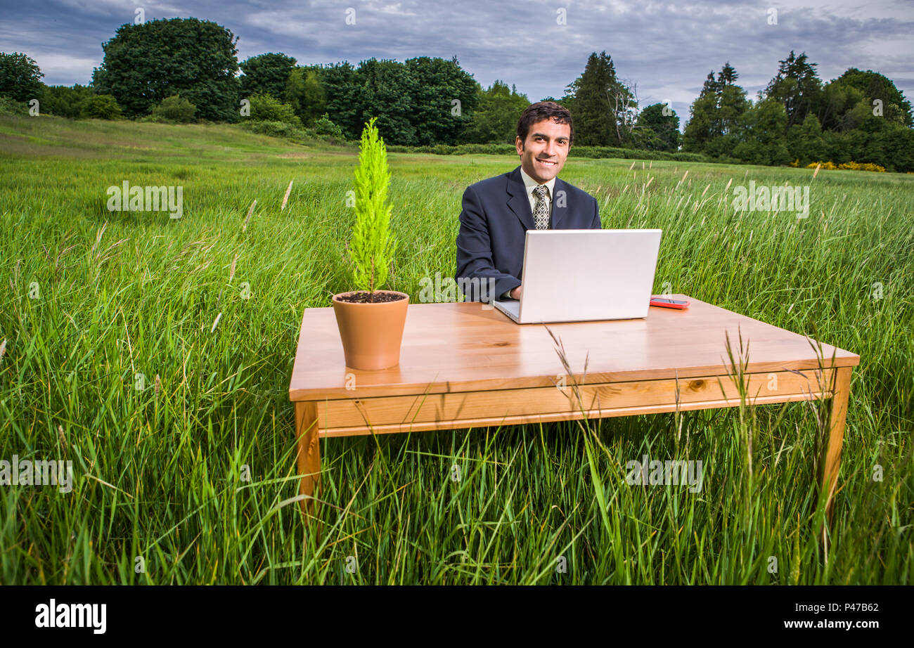 A businessman sitting at a desk with his laptop computer in the middle of a green grassy field. Green business conceptual shot. Stock Photo