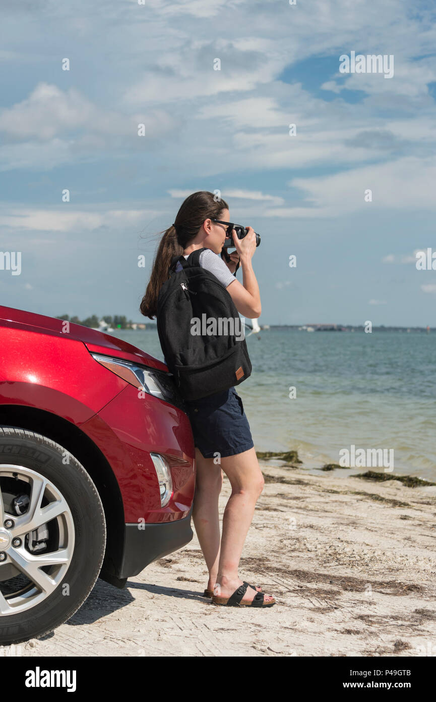 woman tourist leaning against a car taking photos by the sea. Stock Photo