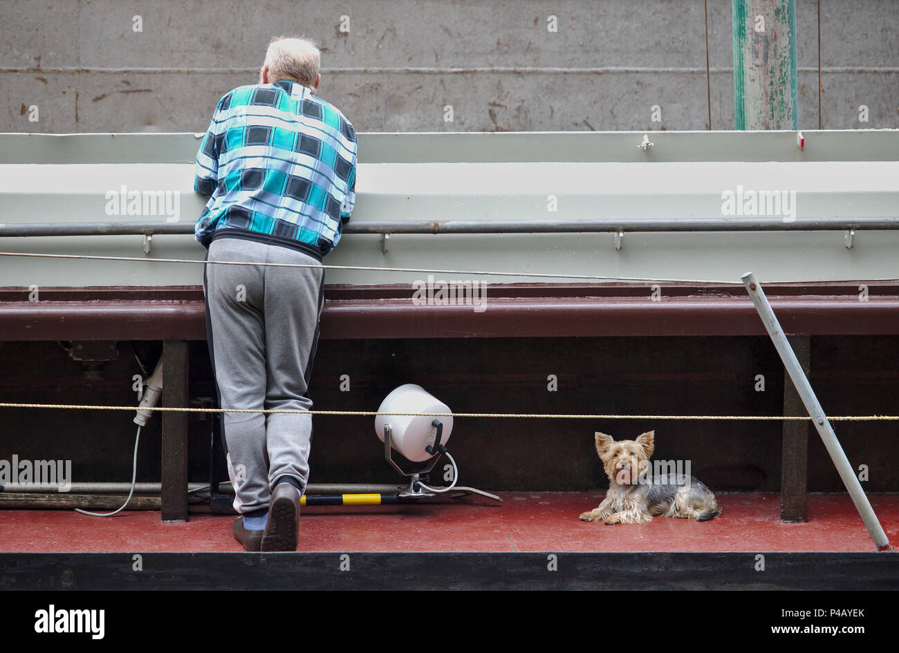Bremen, Germany - June 19th, 2018 - Old man in checkered shirt leaning on a handrail with his back towards the camera with his little dog at his feet Stock Photo