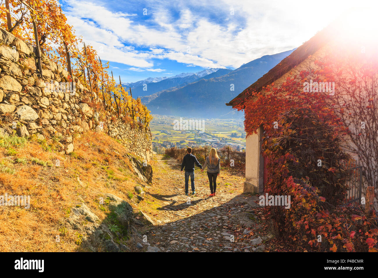 Two people walking on a trail between wineyards and a house cover of american grapes leaves. Poggiridenti, Valtellina, Sondrio province, Lombardy, Italy. Stock Photo