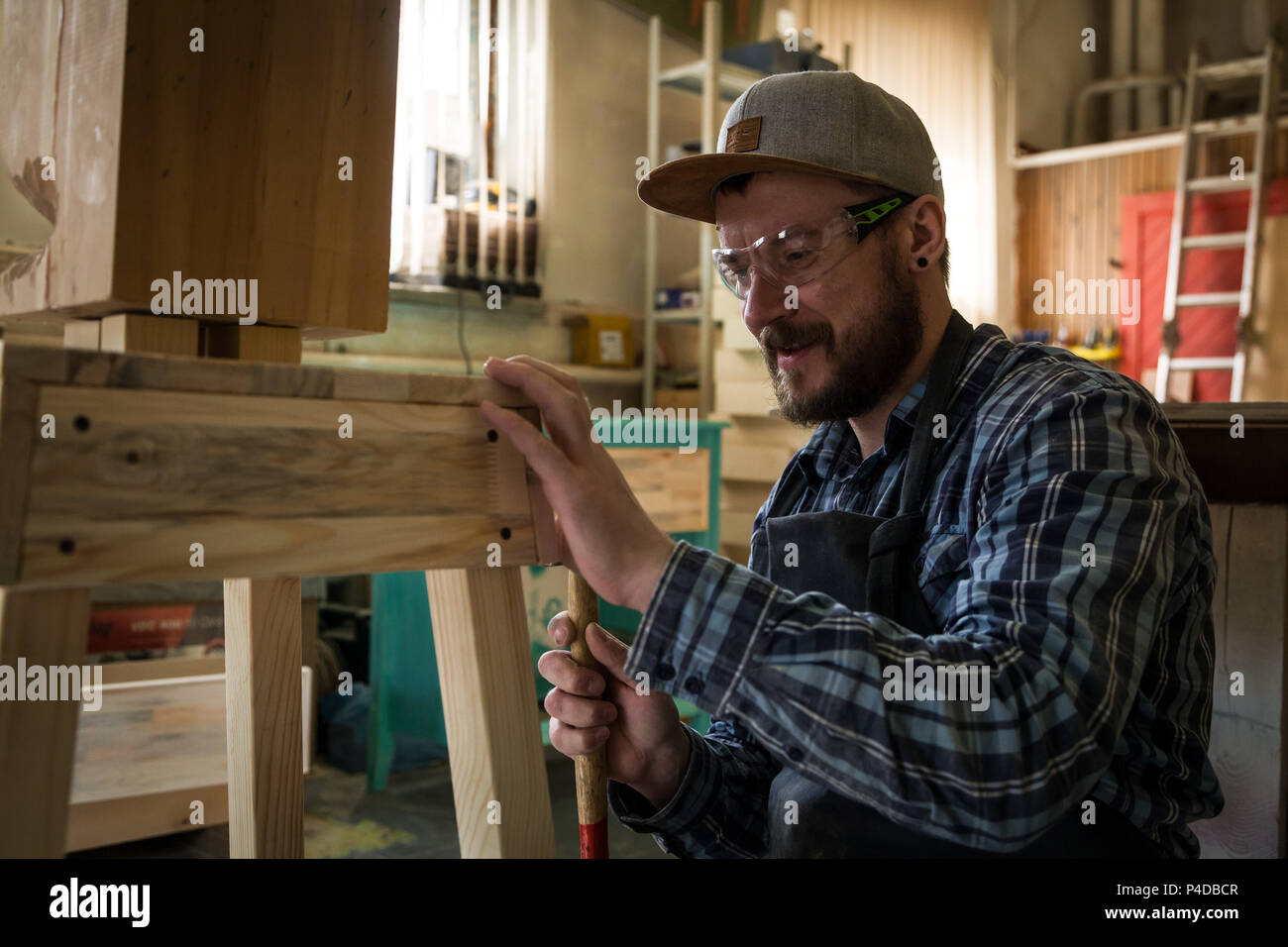 experienced carpenter in work clothes and small buiness owner working in  woodwork workshop, use a hammer to hammer a nail into a stool Stock Photo -  Alamy