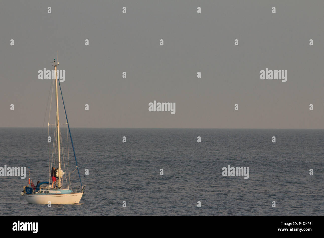 A small blue and white yacht preparing to raise its mainsheet in the early morning light Stock Photo