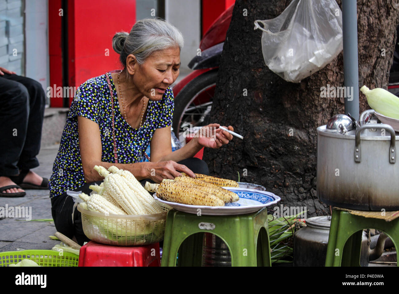 Hanoi, Vietnam - March 15, 2018: Senior lady smoking and selling roasted corn on the street Stock Photo