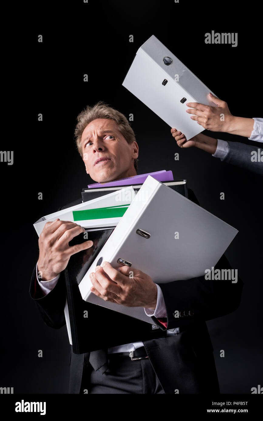 scared overworked mature businessman holding pile of folders isolated on black Stock Photo