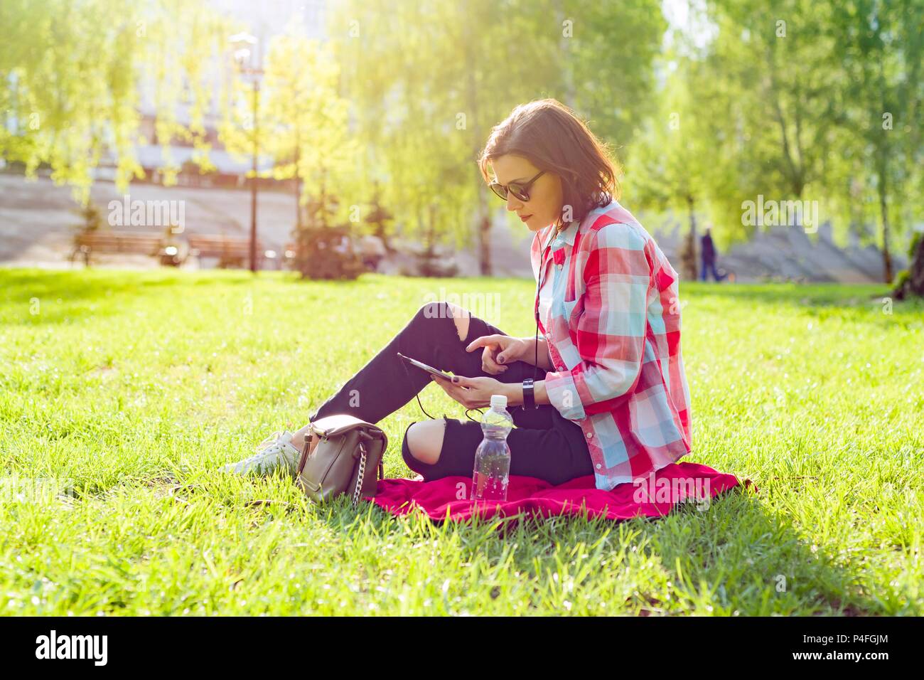 Adult middle-aged woman sits in city park on the grass. In headphones listens audio book, watches video, self-education Stock Photo