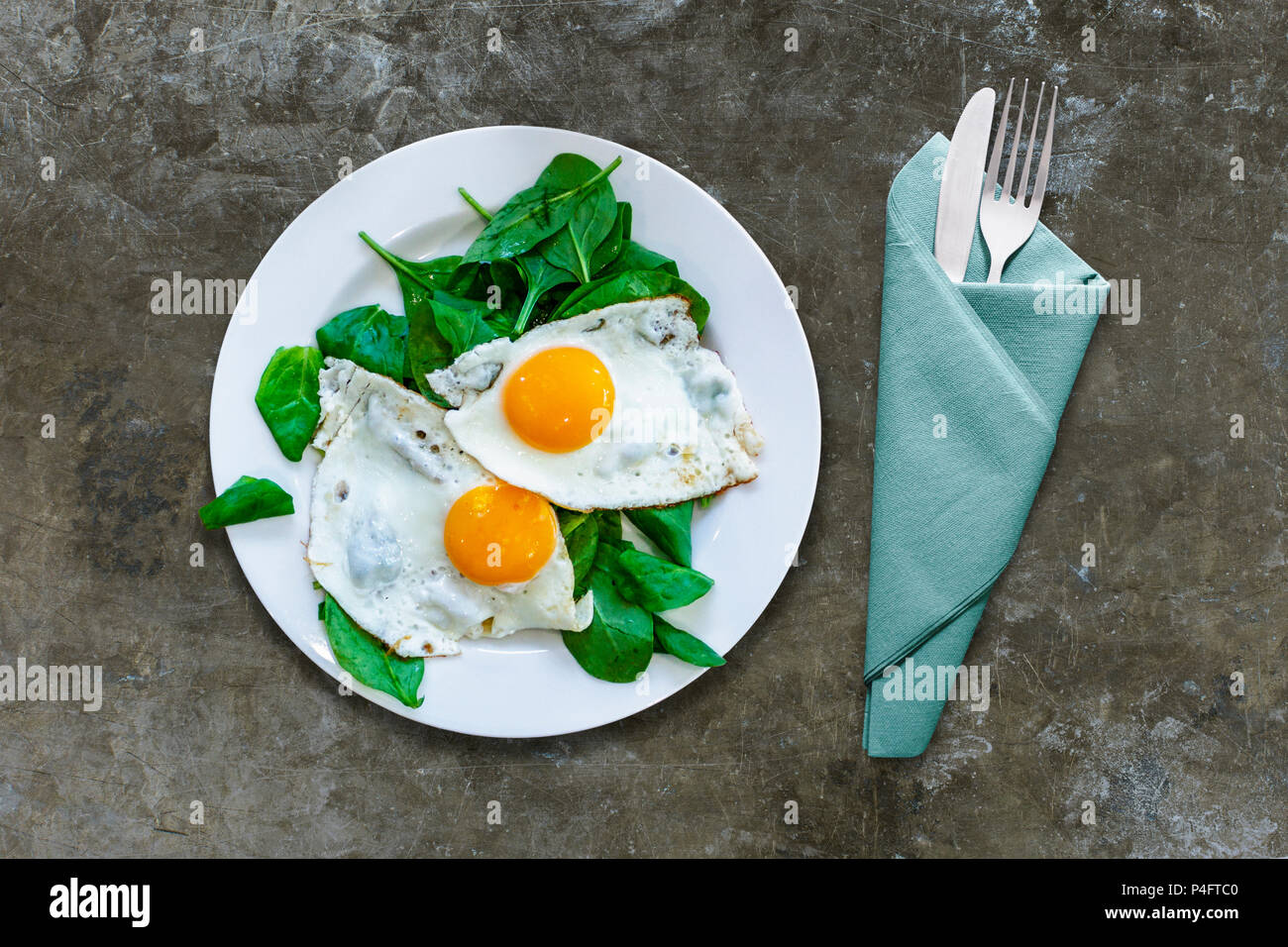 A plate of fried eggs on a bed of spinach with knife and fork wrapped in a napkin on a distressed marble or granite surface Stock Photo