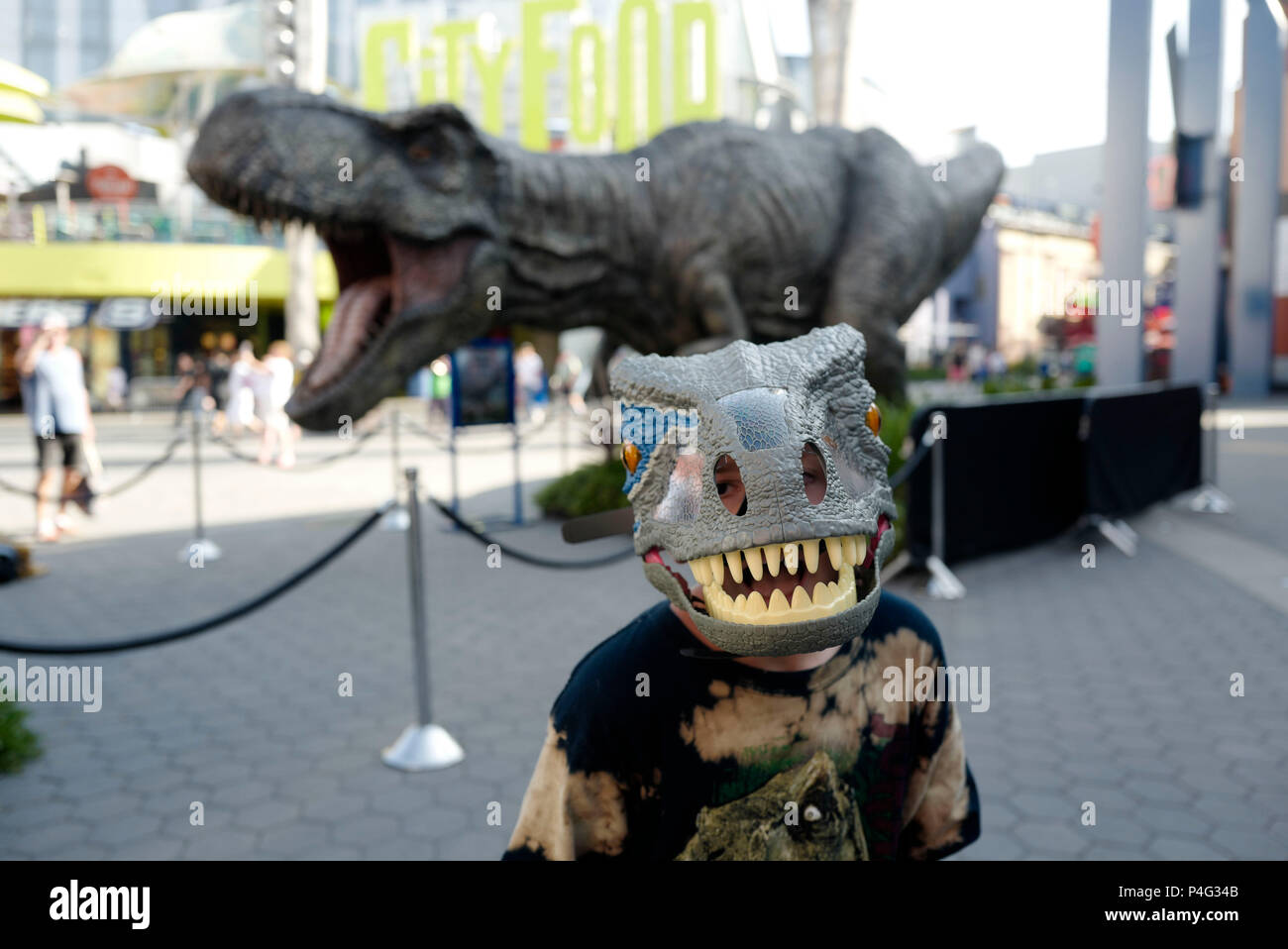 Los Angeles, USA. 21st June, 2018. A boy with a dinosaur mask poses for pictures with a giant Tyrannosaurus rex statue at Universal CityWalk in Los Angeles, the United States, on June 21, 2018. For the promotion of the screening of the new film "Jurassic World: Fallen Kingdom" on Universal Cinema Theatres, a colossal 3-ton Tyrannosaurus rex, a life-sized Gyrosphere original movie prop, as well as original costume, are displayed for public at Universal CityWalk. Credit: Zhao Hanrong/Xinhua/Alamy Live News Stock Photo
