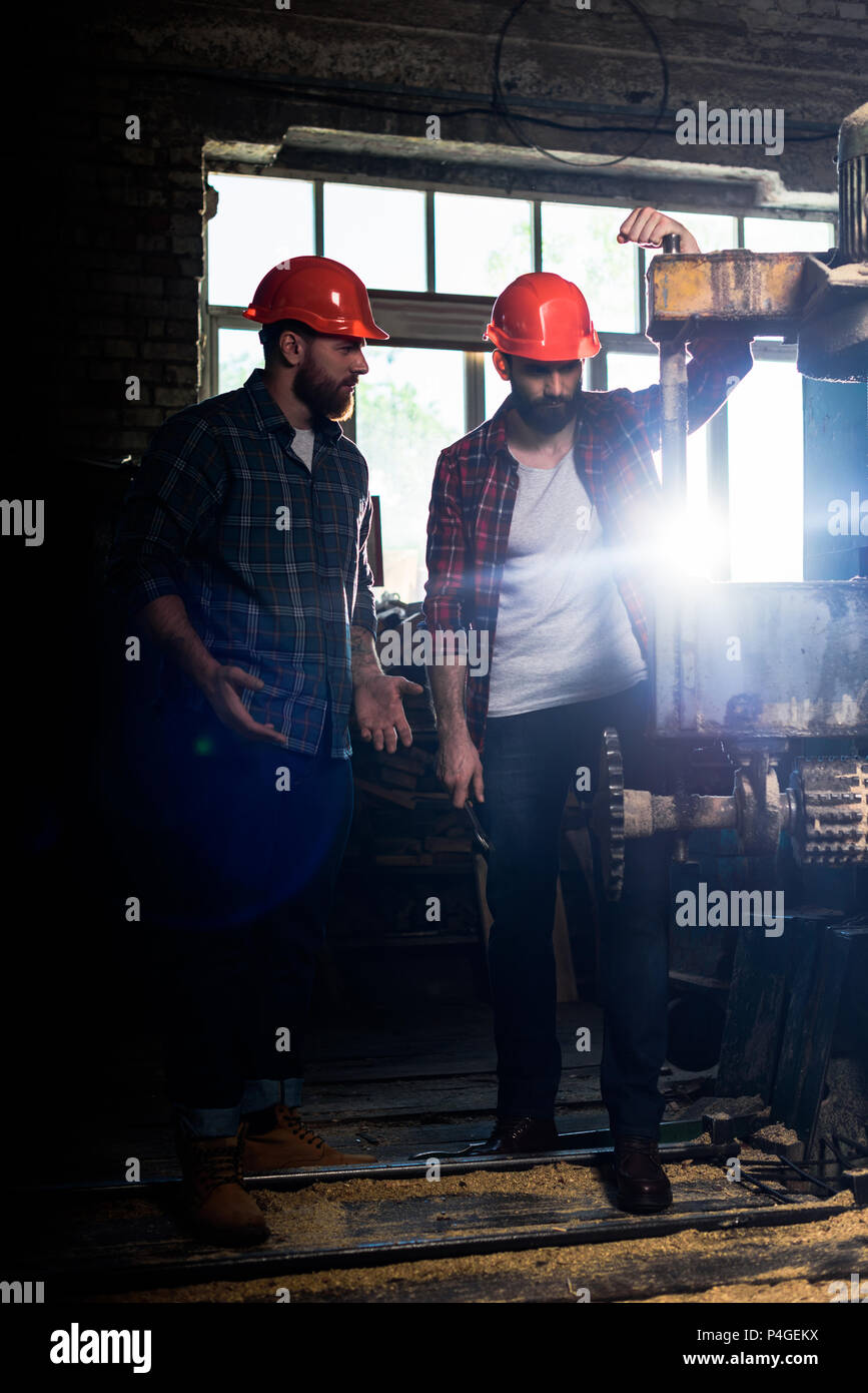 two bearded workers in protective helmets using machine tool at sawmill Stock Photo