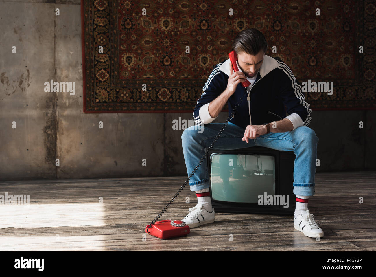 handsome young man in vintage clothes sitting on retro tv set and talking by phone and looking at watch in front of rug hanging on wall Stock Photo