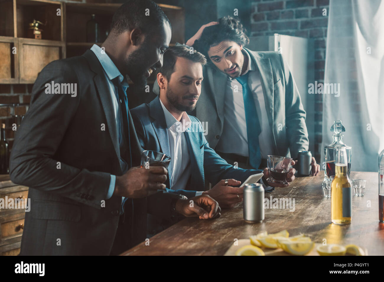 three young men in suits looking at smartphone while drinking whisky together Stock Photo