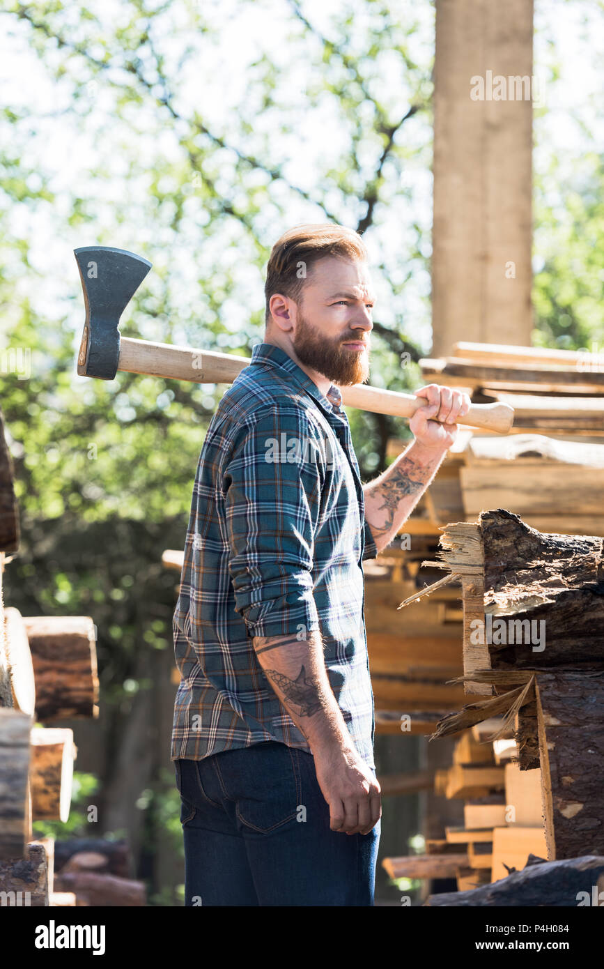 bearded lumberjack in checkered shirt with tattooed hand holding axe on  shoulder at sawmill Stock Photo - Alamy
