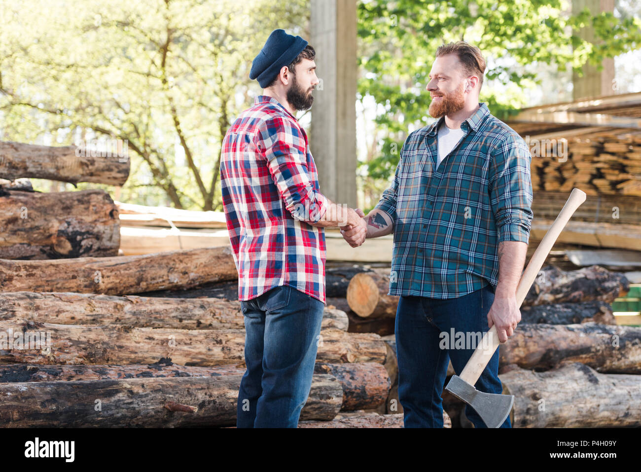 side view of smiling lumberjack with tattooed hand holding axe and shaking hands with partner at