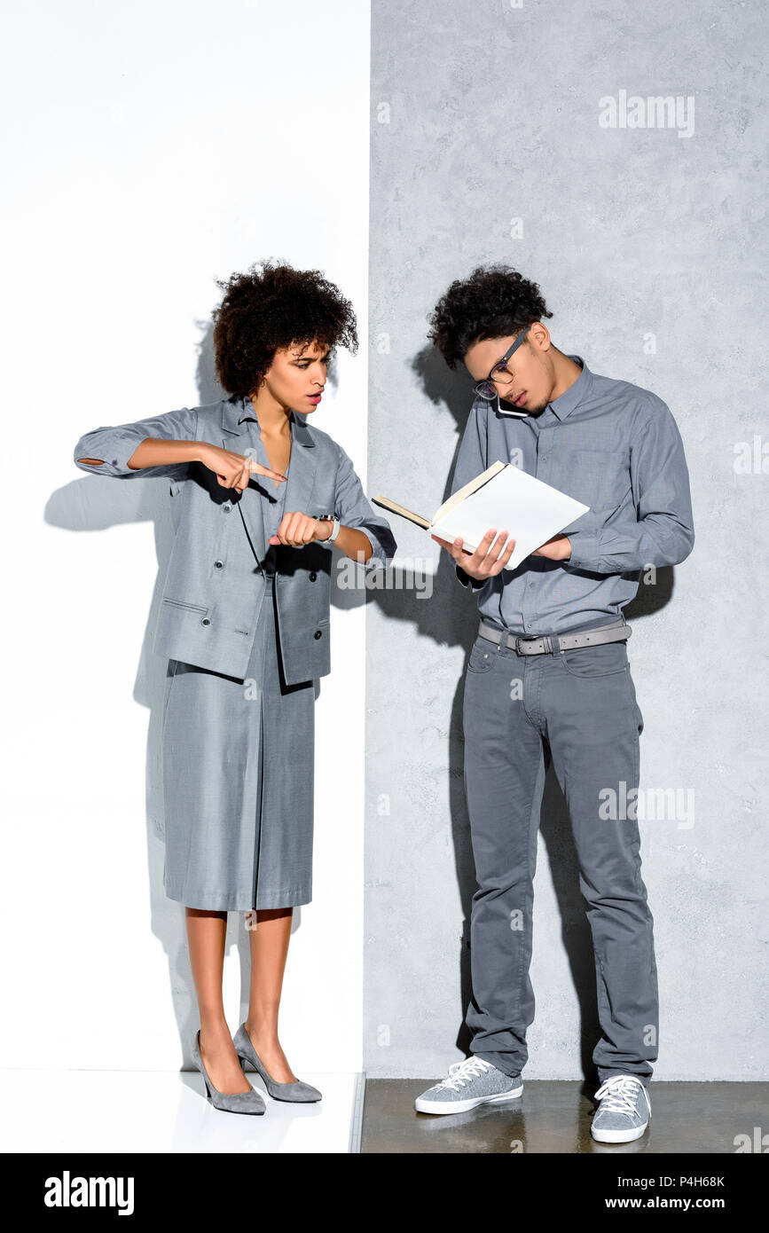Young african amercian businessman holding folder and talking on phone while angry businesswoman pointing on watch on grey and white background Stock Photo