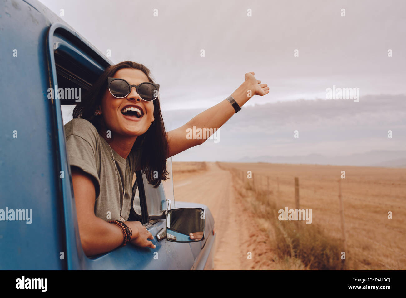 Happy woman out on a road trip in a car. Young female raising her hand out of the car. Enjoying on countryside road trip. Stock Photo
