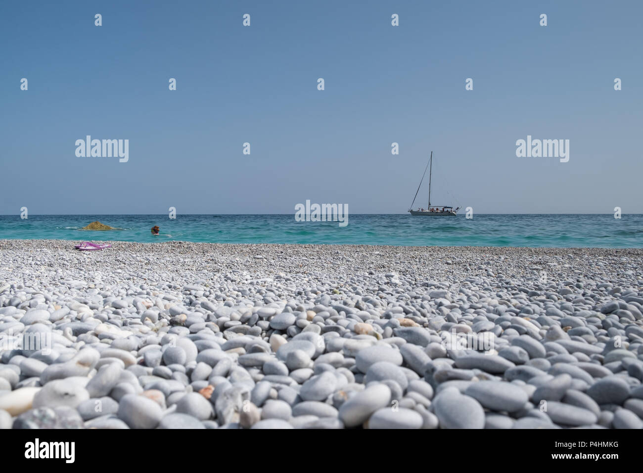 Beautiful pebble beach with a sailing boat and swimmer in the bacground. Stock Photo