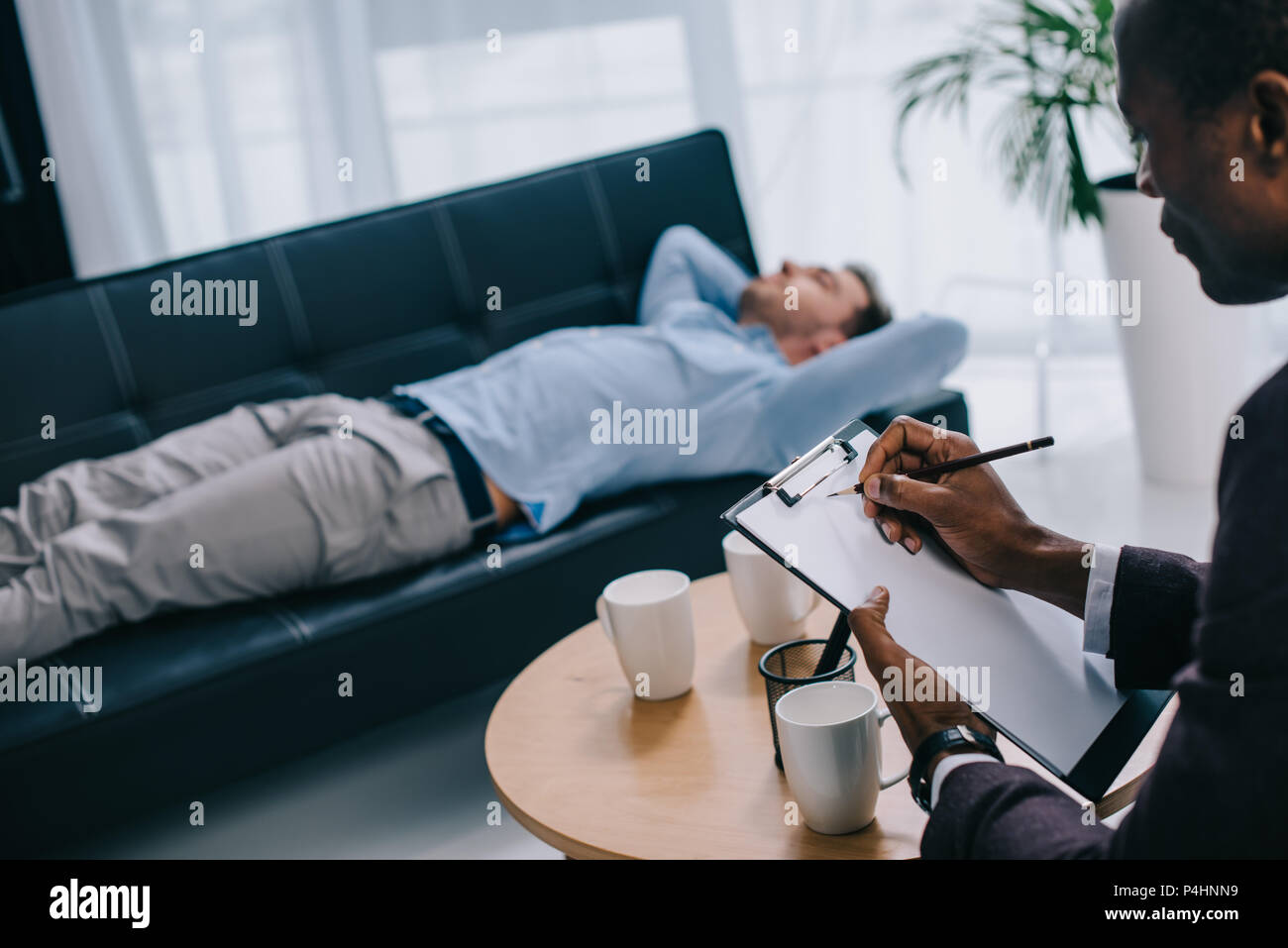 Counselor writing in clipboard and young male patient laying on sofa Stock Photo