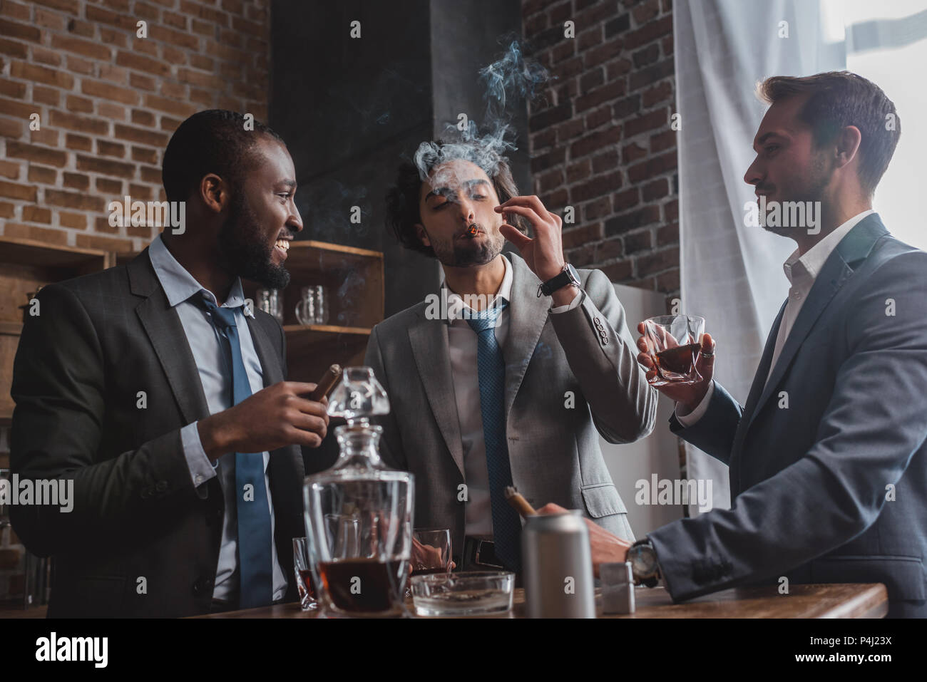 multiethnic male friends in suits smoking cigars, drinking whiskey and talking Stock Photo
