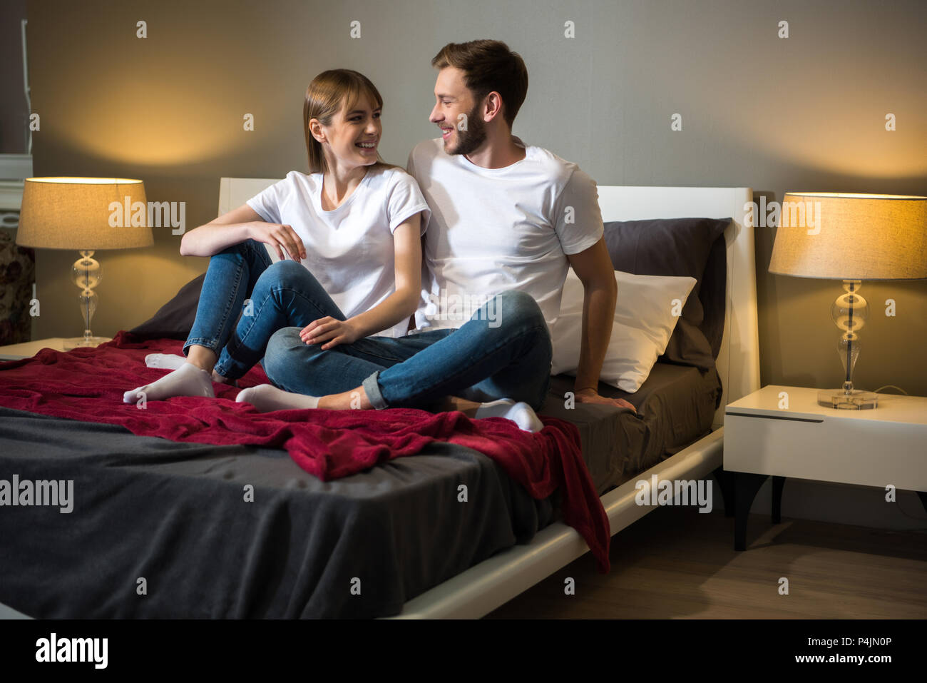 Happy Young Couple Sitting On Bed In Cozy Modern Bedroom