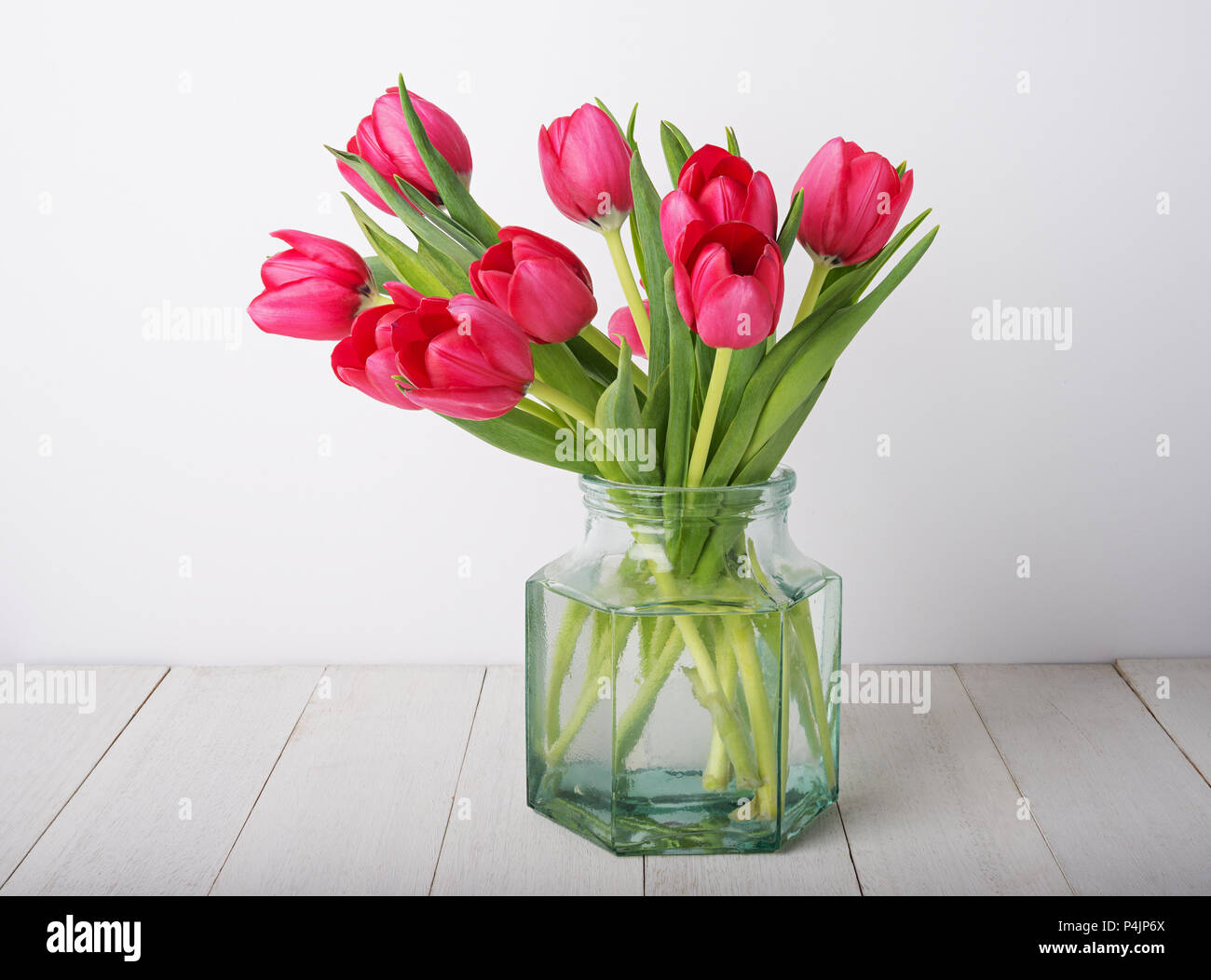 bunch of red tulip flowers  in a glass vintage jar on rustic wooden table against white plaster wall Stock Photo