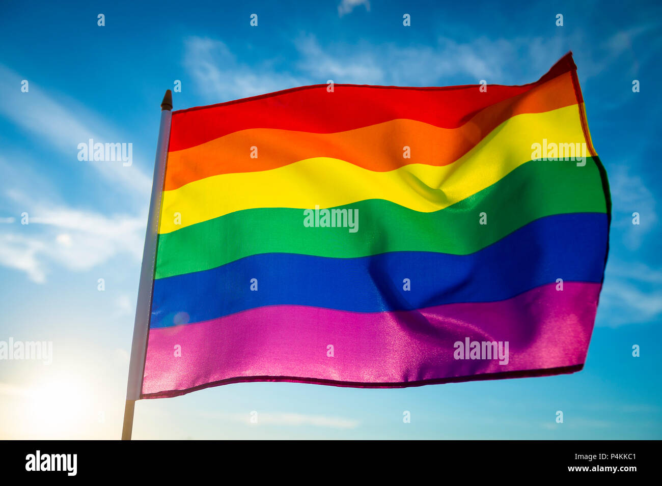 Backlit gay pride rainbow flag flutters in the breeze with lens flare. Stock Photo