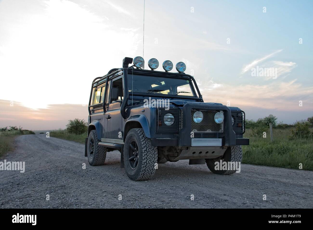 A blue off road Land Rover Defender 90, County on grey track with sunset behind. Stock Photo