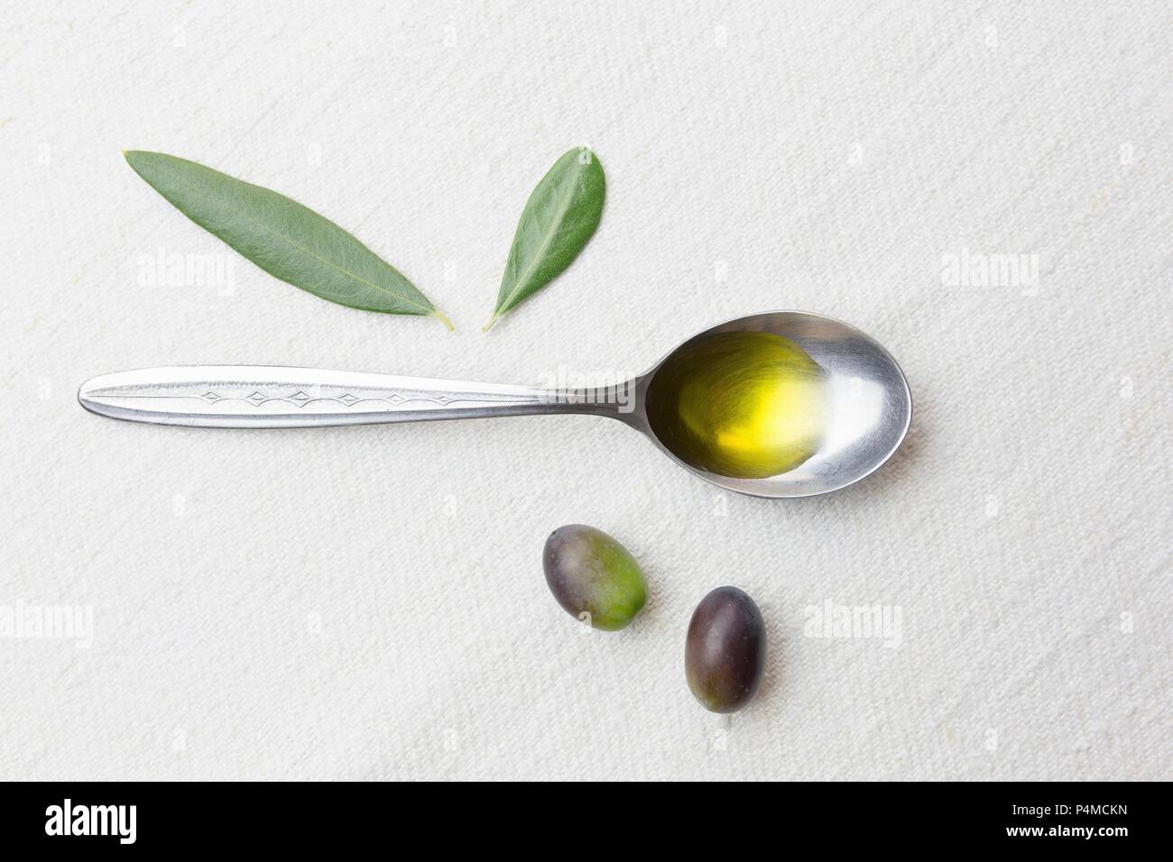 A spoonful of olive oil, olives and olive leaves on a white surface