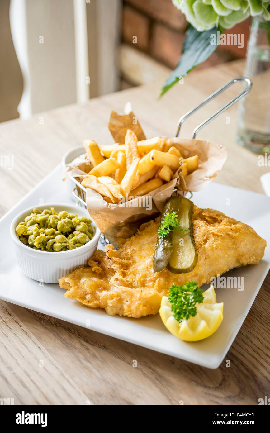Gourmet fish and chips deep fried cod fillet in a beer batter with chips in a basket, gherkins pickle, mushy peas and half a lemon Stock Photo
