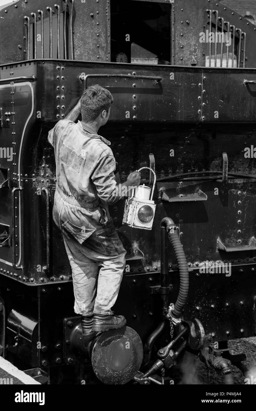 Portrait, black & white rear view of steam train driver in dirty overalls, from behind, climbing aboard vintage UK steam locomotive to change lamp. Stock Photo