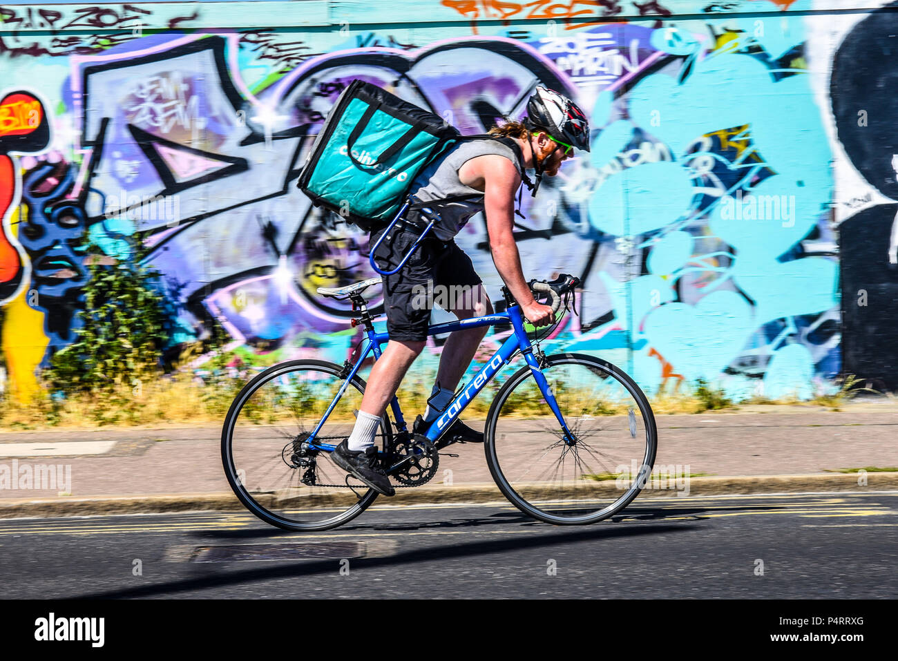 Deliveroo bicycle delivery rider struggling in the heat, riding past bright graffiti in Southend on Sea, Essex, UK. Cyclist take away food delivery Stock Photo