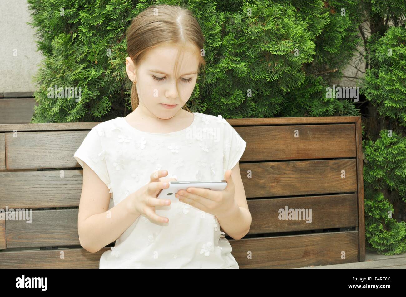 Child, girl 7-9, blond hair, standing on a bench and playing on smartphone, outdoor, retro style, front view Stock Photo