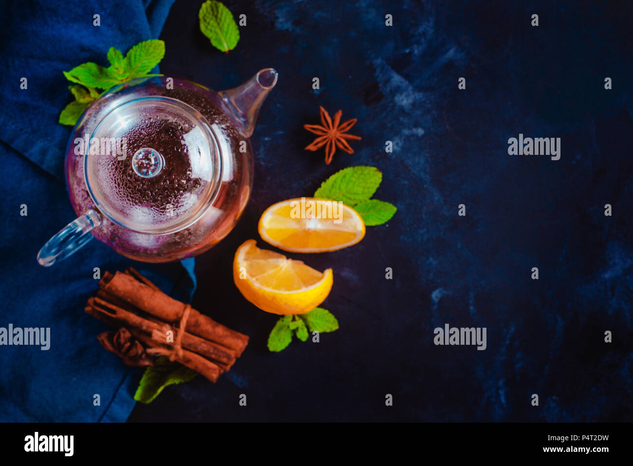 Tiny glass teapot with lemon slices, cinnamon and mint leaves on a dark background. Vibrant colors hot drink still life with copy space. Tea time flat Stock Photo