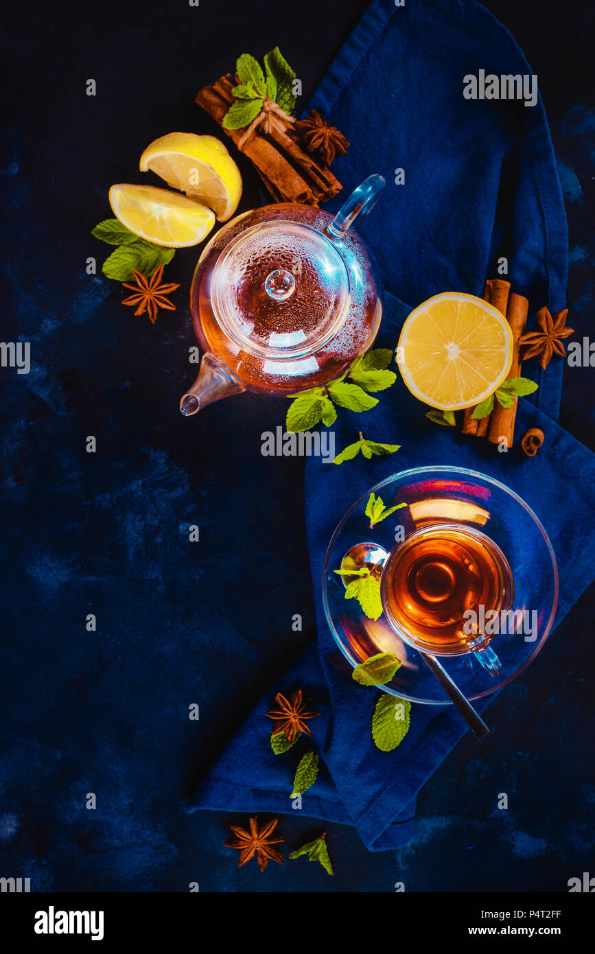 Tea cup and a tiny teapot with lemon slices and mint leaves on a dark background. Vibrant colors hot drink still life with copy space. Tea time flat l Stock Photo
