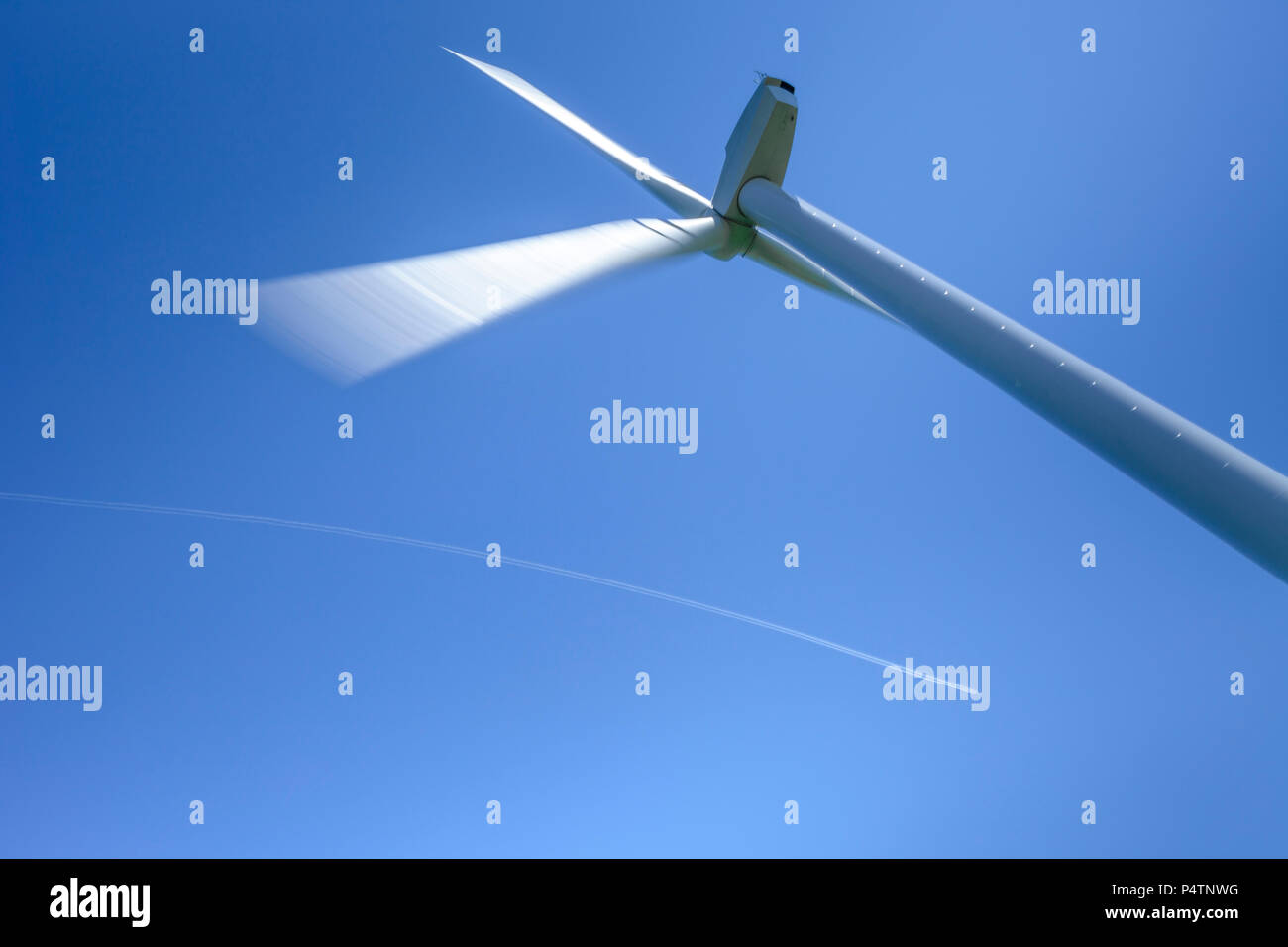 Wind turbine in action in a windmill park against a clear blue sky, an airplane with contrails