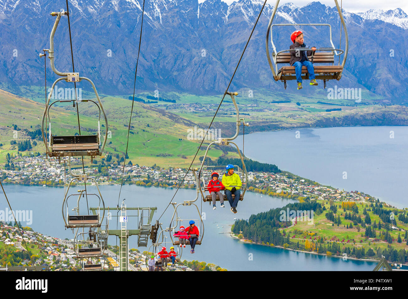 Queenstown, NZ – April 15, 2018: Fun riders with their carts on a cable chair lift going up the hill top for a luge ride with views of Lake and town. Stock Photo
