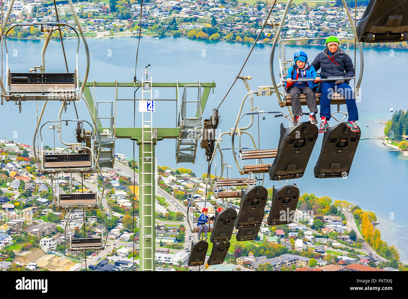 Queenstown, NZ – April 15, 2018: Fun riders with their carts on a cable chair lift going up the hill top for a luge ride with views of Lake and town. Stock Photo