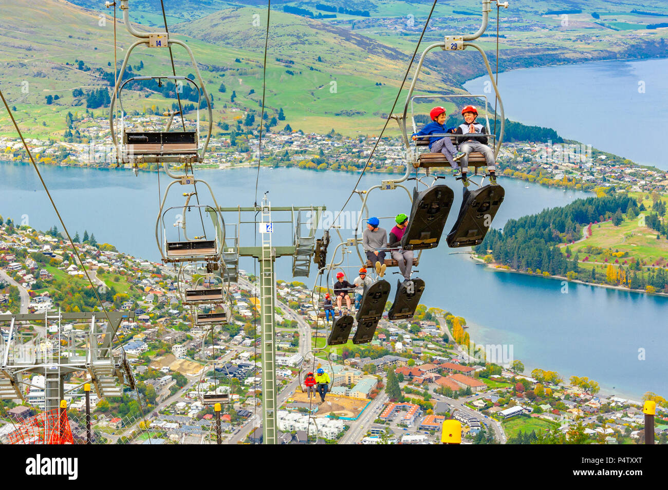 Queenstown, NZ – April 15, 2018: Fun riders with their carts on a cable chair lift going up the hill top for a luge ride with views of Lake and town. Stock Photo