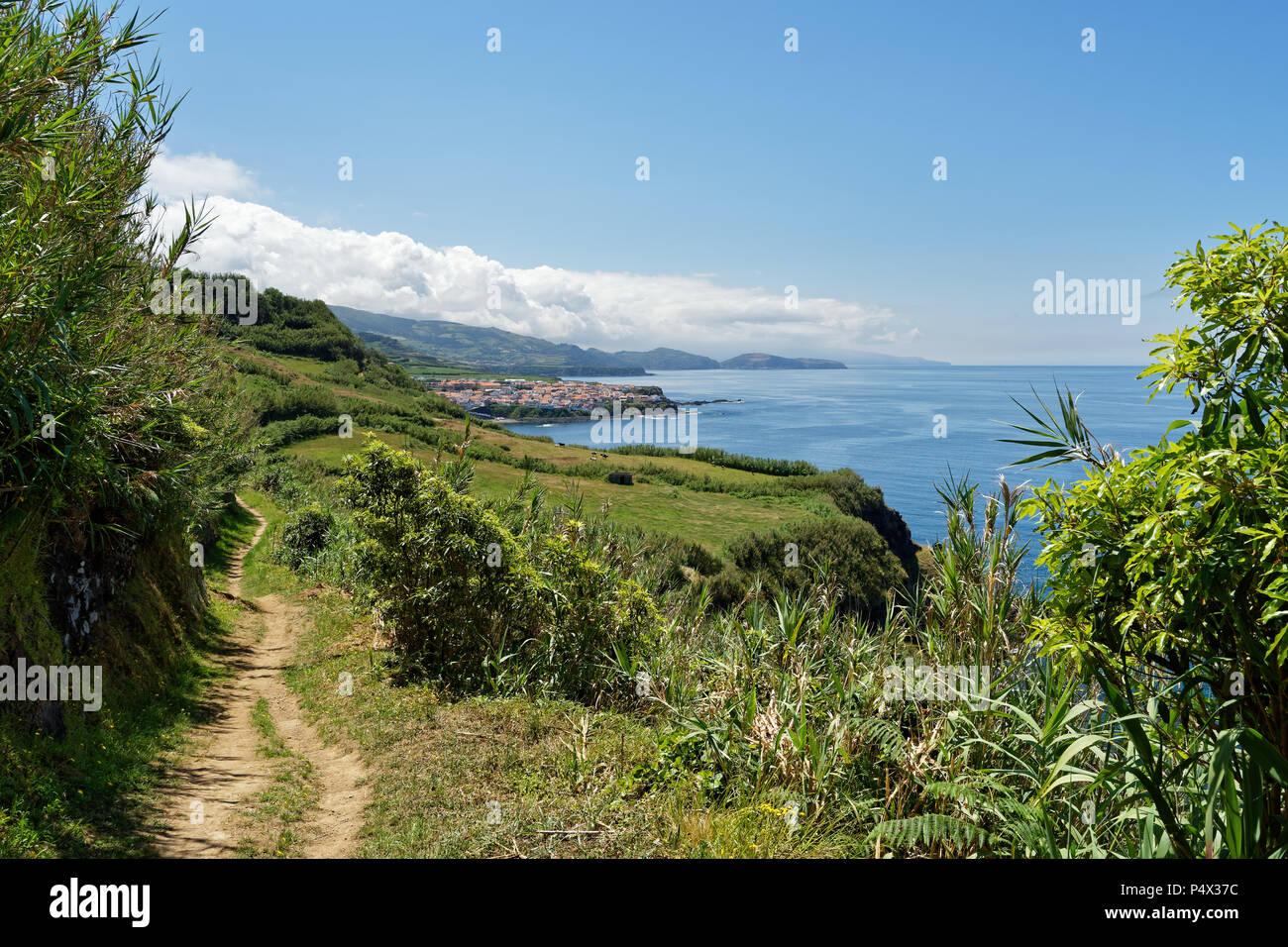 Coastal walk on the Azores island of Sao Miguel in sunny weather, the view follows the trail past pastures to a village and mountains with clouds - Lo Stock Photo