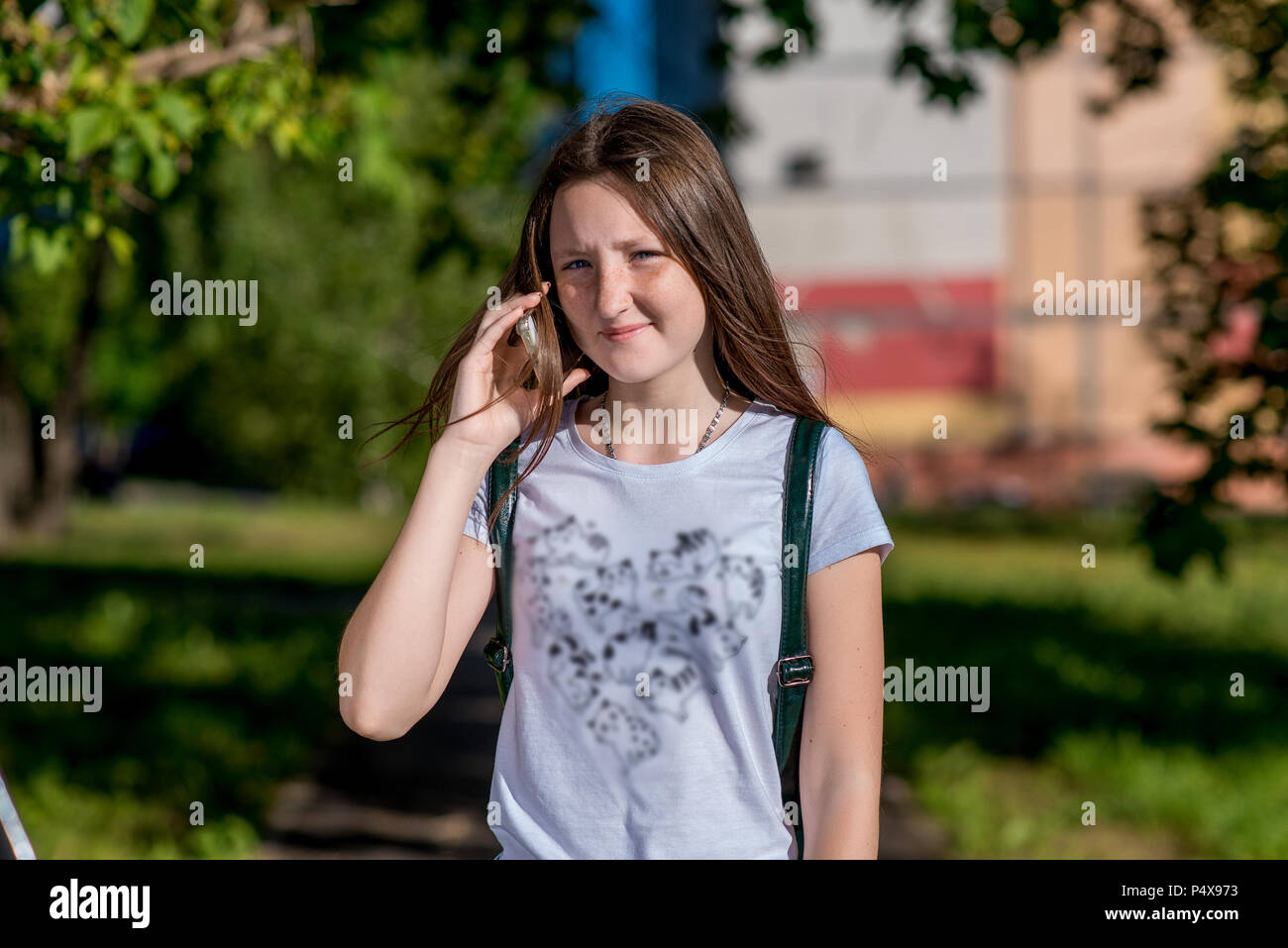 Girl schoolgirl. Summer in nature. Holds the hands of a smartphone. She calls on phone. Smiles happily. In the summer In the bright sun. In a white T-shirt behind his backpack. Stock Photo