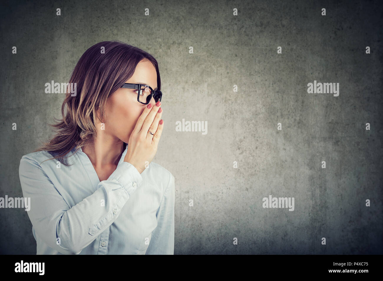 Young business woman covering mouth while telling a secret posing on gray wall background. Stock Photo