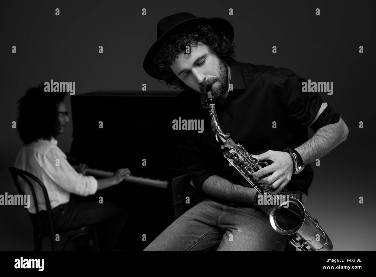 black and white shot of man playing saxophone while his partner playing piano blurred on background Stock Photo