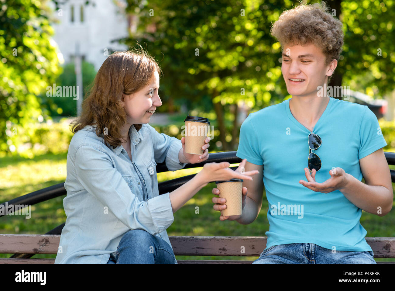 A guy with a happy girl in a park on nature. In summer they sit on bench. He holds cups with coffee or tea in his hands. He gestures emotionally with his hands. Stock Photo