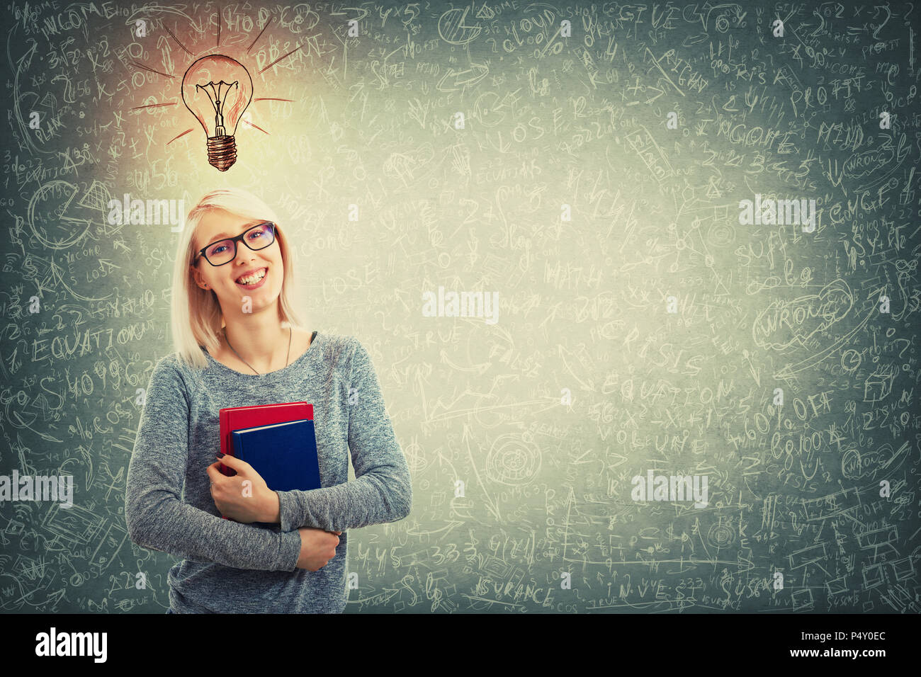 Young woman wearing glasses and holding two books stand over blackboard background with a light bulb sketch above head, solving sophisticated mathemat Stock Photo