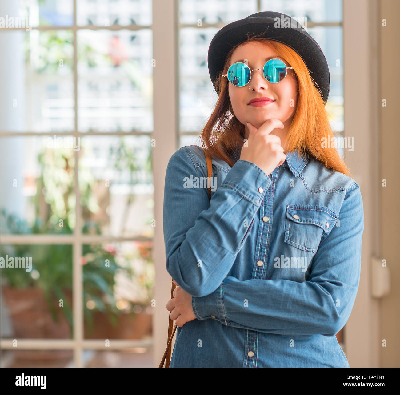 Stylish redhead woman wearing bowler hat and sunglasses looking confident at the camera with smile with crossed arms and hand raised on chin. Thinking Stock Photo