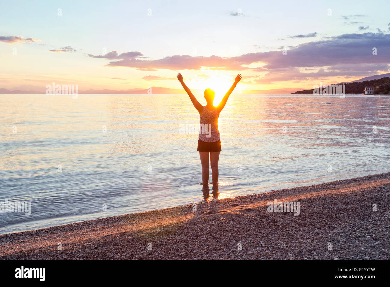 Greece, Pelion, Pagasetic Gulf, woman on the beach with raised arms at sunset, Kalamos in the background Stock Photo