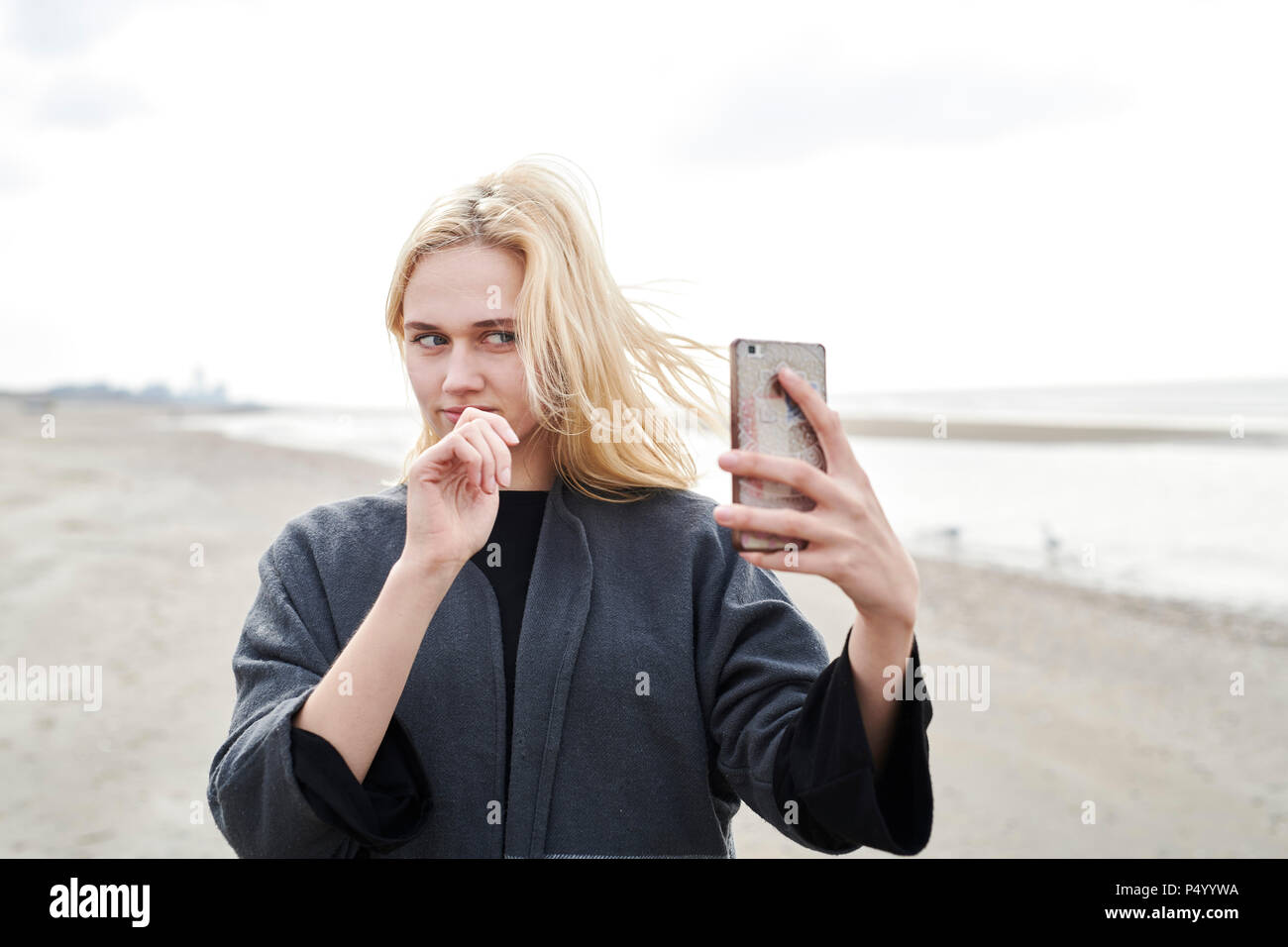 Netherlands, portrait of blond young woman taking selfie with smartphone on the beach Stock Photo