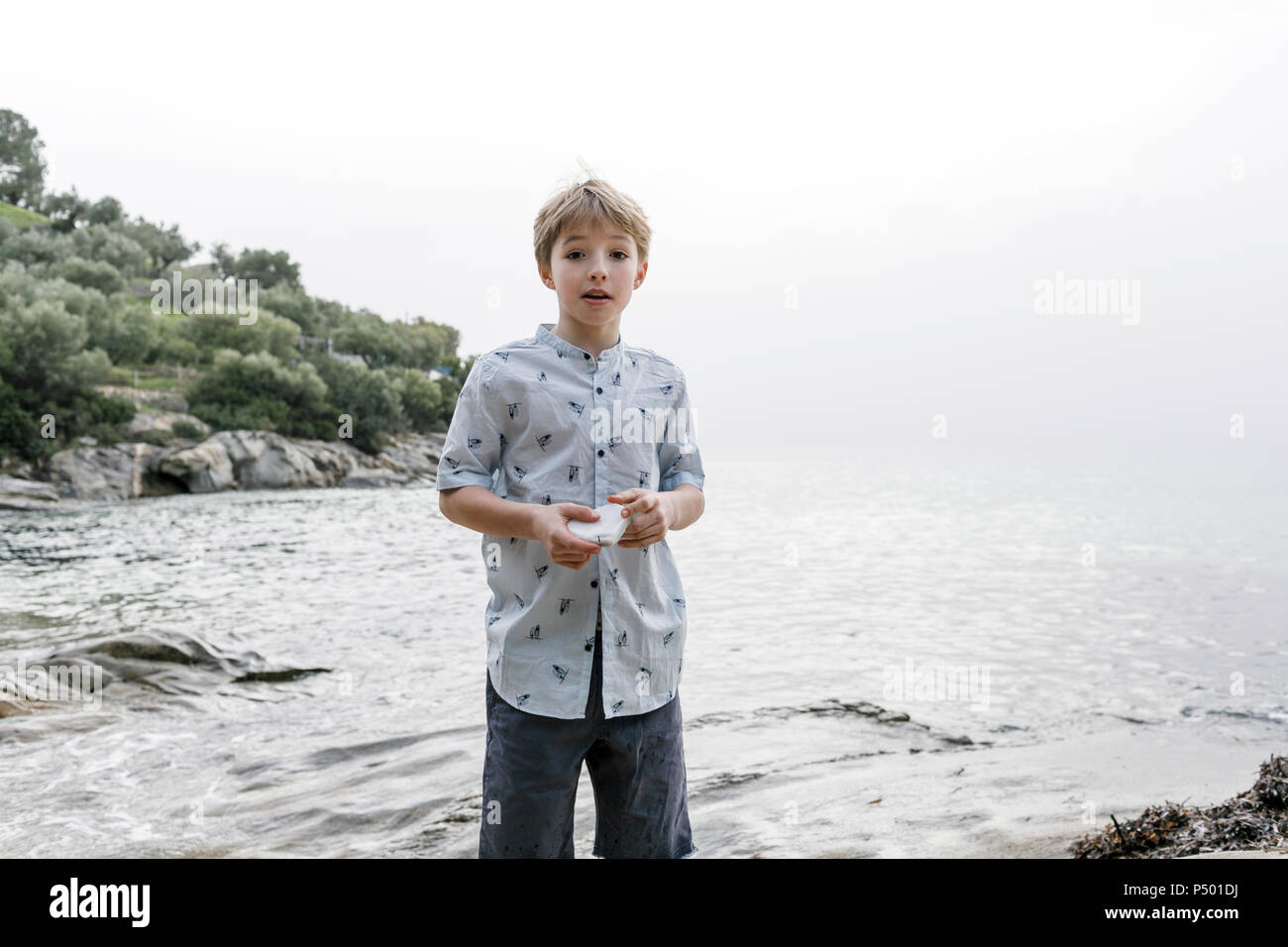 Greece, Chalkidiki, portrait of blond boy standing in front of the sea holding stone Stock Photo